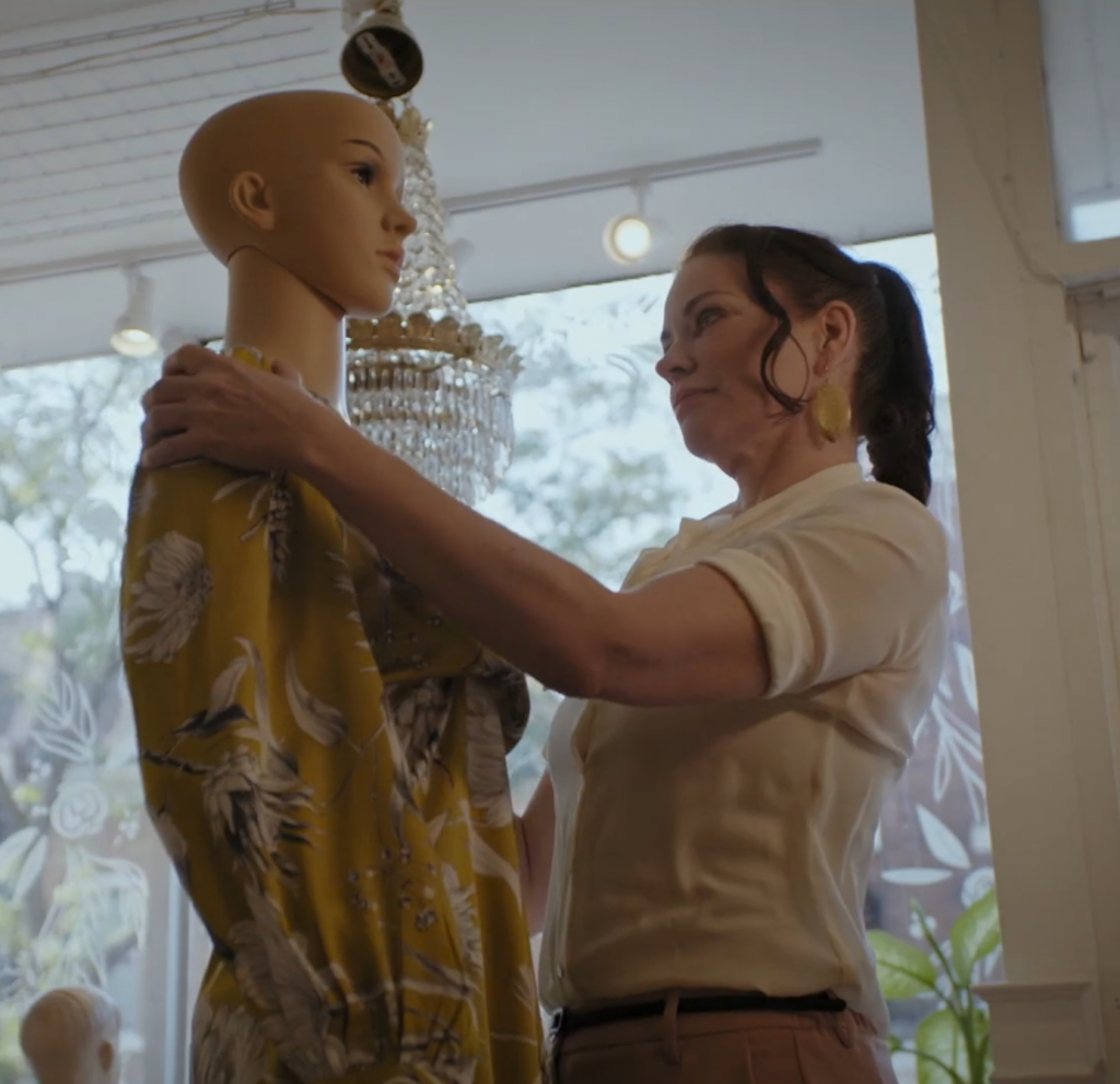 A woman wearing white holding up a mannequin in a dated yellow dress. 
