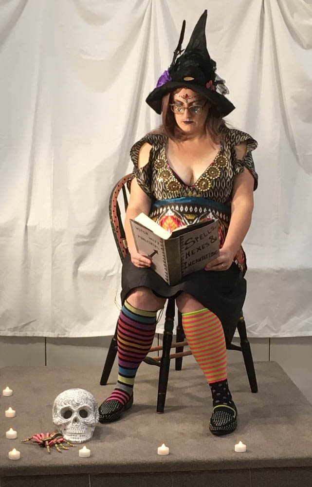 a witch sits reading her book Speels, Hexes & Incantations by Witch Way Publishing, wearing her patterned finery surrounded by candles, a voodoo skull and a pink and black spider