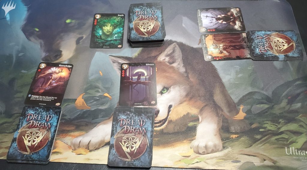 A photograph of a three-player game in action. The player furthest to the left has played Knowledge, a card with 5 strength, 1 damage, and the rules text "Summon: You may discard the top card of the deck. If it's level 6 or greater, you may summon it." The middle player has played Silence, a card with 9 strength and 3 damage. The player to the right has played The Guardian, with 4 strength and 3 damage, and Betrayal, with 6 strength and 3 damage. In the middle of the table is the main deck, and to its left is the discard pile. The discard pile contains the card Earth, with 2 strength and 3 damage.