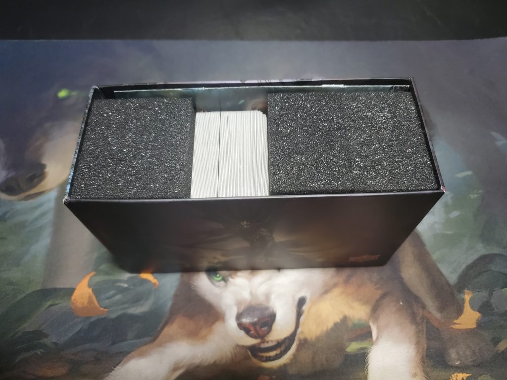 A photograph of the game's components while the game is still in the box. From left to right there is a foam block, a one hundred card deck, and two more foam blocks. Each foam block is roughly the same size as the card deck. There is a thin rule book in a small gap above the components.