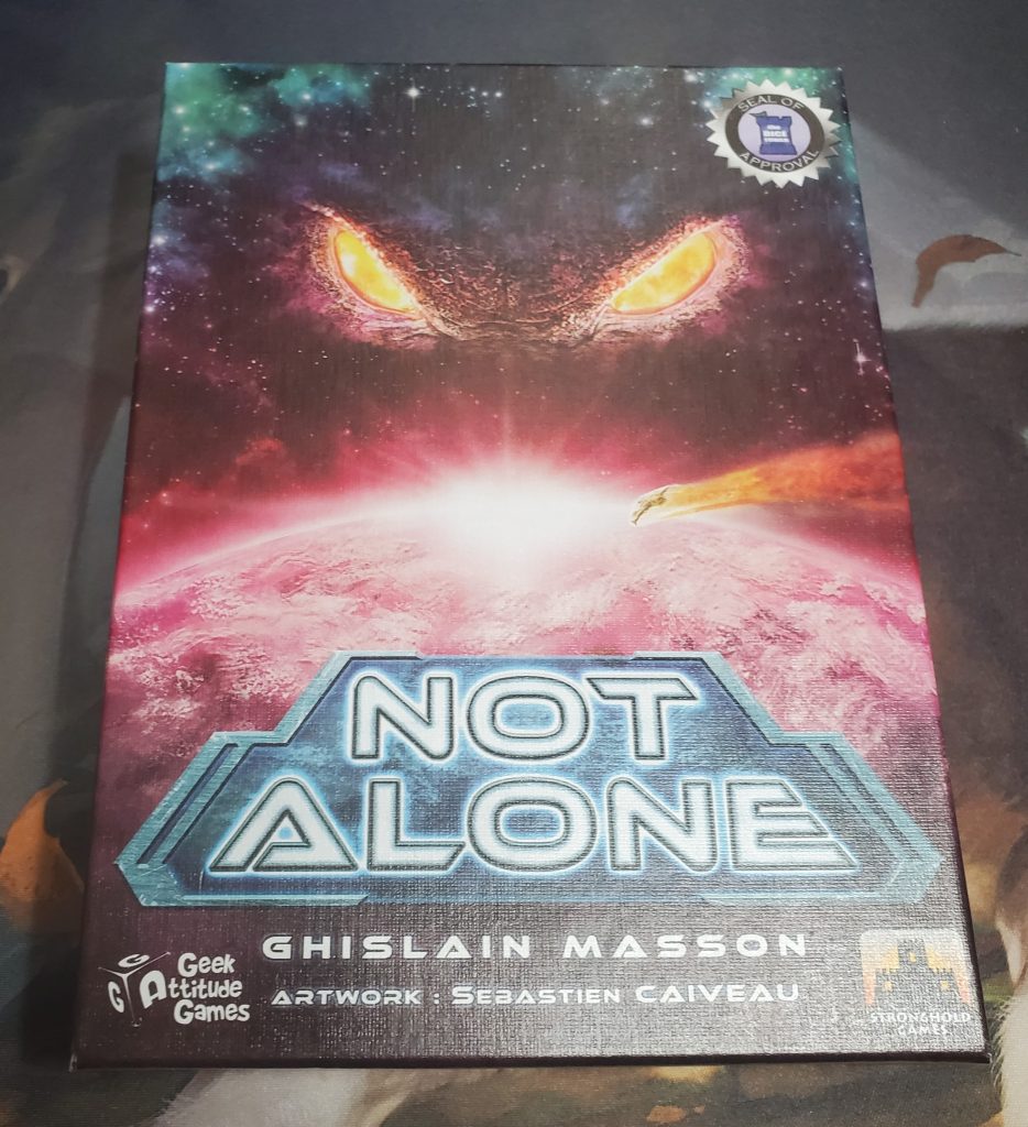 A photograph of the Not Alone game box. The art depicts a small ship on a collision course with a pink planet, and glowing yellow eyes in the galaxy above. In the top right corner is the Dice Tower Seal of Approval. At the bottom is the text "Not Alone" in white in a steel grey box. Underneath that are the names of the designer, Ghislain Masson, and the artist, Sebastien Caiveau. In the left corner is the Geek Attitude Games logo and in the right is the Stronghold Games logo.