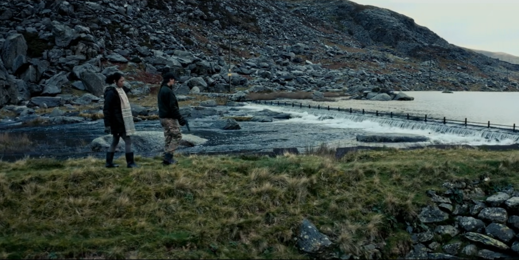 A photograph from the film showing two characters walking outdoors. They are passing by a river in front of a mountain.