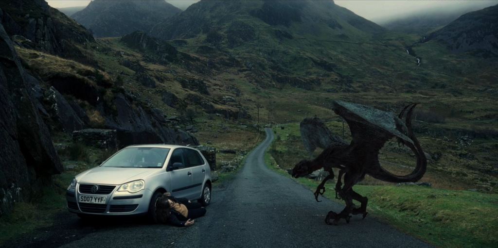A photograph from the film. A road leads into the mountains. On the left side of the road is a white car and a woman lying on the ground. On the right is a CGI dragon.