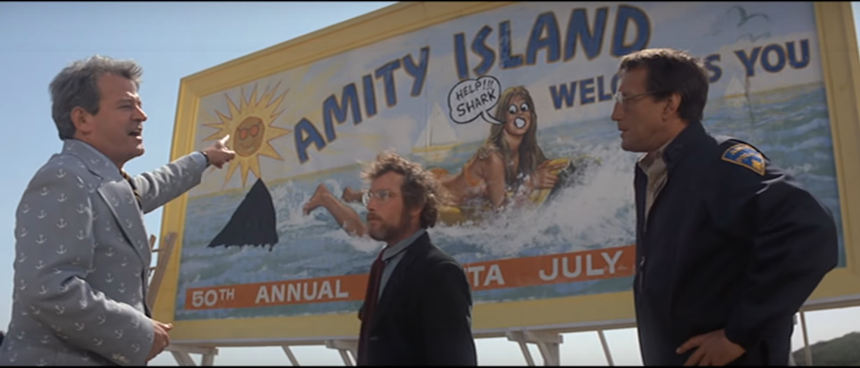 The mayor of Amity angrily points at a billboard that has been vandalized with a drawing of a shark's fin. Brody and Hooper listen in exasperation.