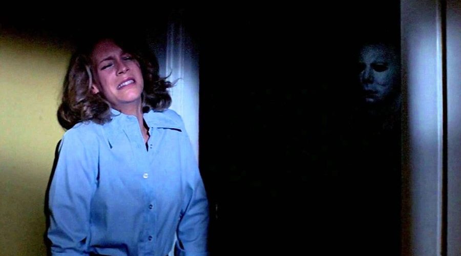 Laurie Strode is against the wall scared while Michael Meyers is in the background