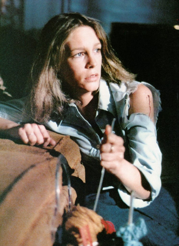 Laurie Strode is sitting down hiding from Michael Meyers, holding a knitting kneedle.