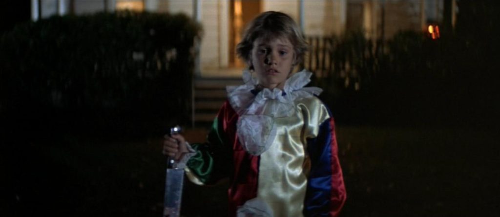 A young boy revealed to be Michael Meyers is standing outdoors in a clown costume holding a kitchen knife