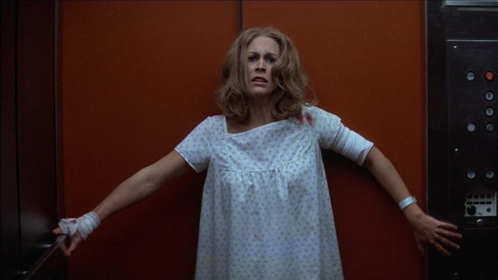 Laurie Strode is leaning against an elevator door. She's wearing a hospital gown, looking scared.