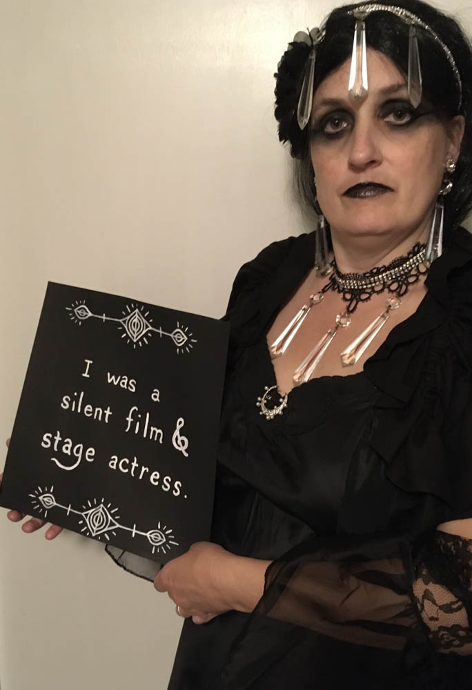 Jennifer Weigel in black tragic Gothic dress with crescent moon pendant & crystals and dark, kohl outlined eyes holding sign, from wax museum performance