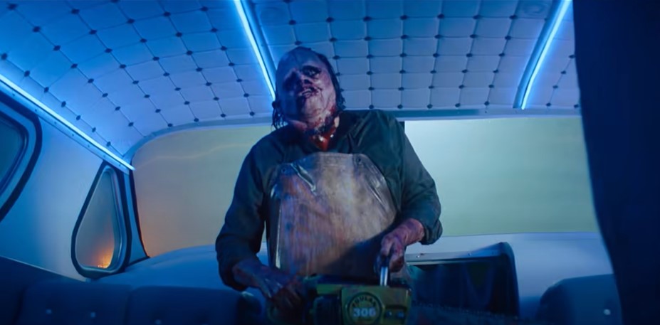 Leatherface enclosed in a bus wearing his bloody face mask. Streaks of red running down his chin, a grimy mustard yellow apron covers his chest, while he grips his rusted chainsaw in hands. The white-blue neon lights illuminating within the bus. 