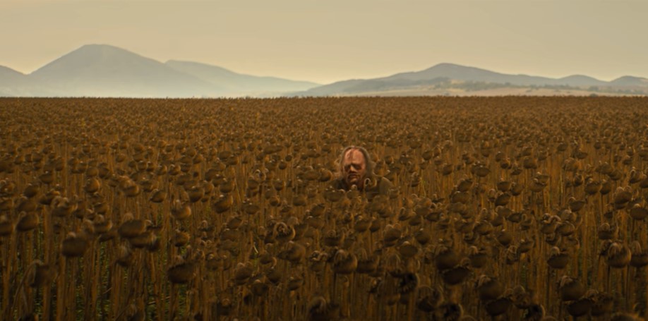 Image of Leatherface standing in a field wearing a human face mask. Dark browns mixed with hues of grey skies adding chilling atmosphere. Mountains are seen in the distance adding scale to Leatherface's stature. 