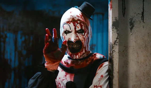 David Howard Thornton in white and black clown makeup splattered in bright red blood. Dressed as Art the Clow with a sinister smile, waving hello. Image from Terrfier 2. 