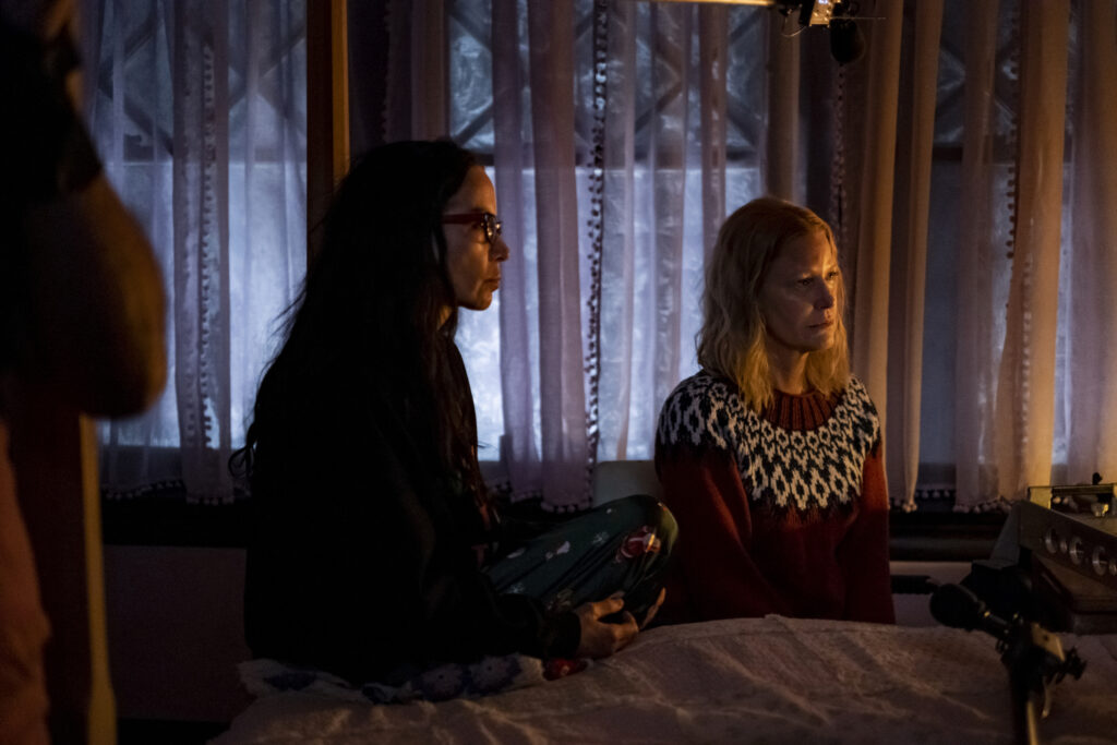 Gretchen and Darla sit next to each other staring at someone in a bed. White opaque curtains shield the windows behind them as the room they sit in sits in faint lamp luminescence. Grethen, sitting on the left wear glasses and is wearing a black long sleeve shirt, while her friend Darla sits to her left in a red and black sweater with a white diamond pattern stitching. Her hair is medium length blonde, resting just past the base of her neck.