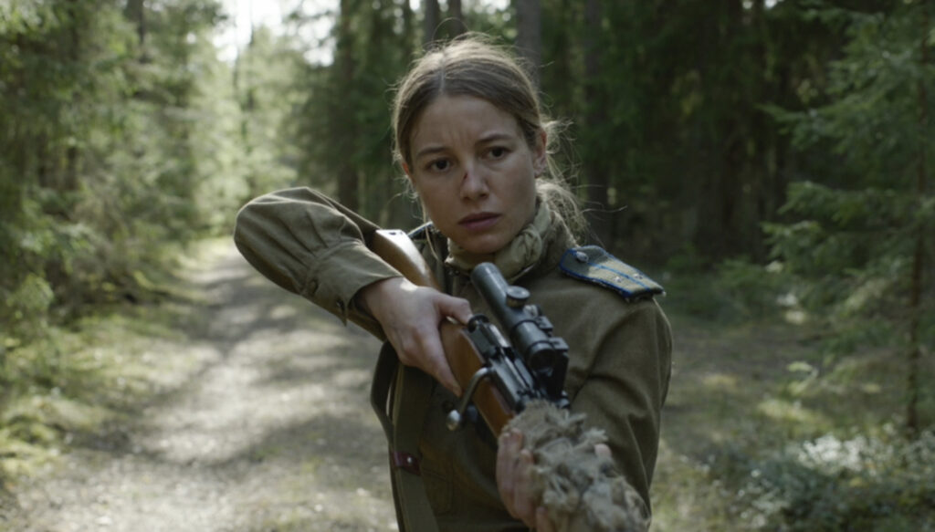 Actress Charlotte Vega is seen holding her rifle in a cautionary firing position draped in her military garbs during day time. Her character is walking along a gravel road deep in a forest on the outskirts of town. Her brown hair is pulled in a pony tail and a small cut across the bridge of her nose. 