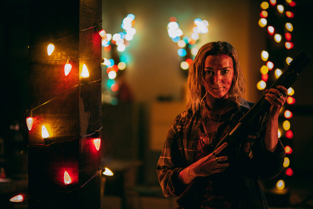 Riley Dandy as Tori Toom wielding a pump action shot gun. Her blonde hair drapes down the side of her blood-smeared face as orange, red, white, and green christmas lights twinkle around her. Each hand has 3 rings on her fingers (1 on her index, the other on her middle, and the last on her ring finger). She is draped in a loose buttoned up long-sleeve black, grey and white plaid shirt. 