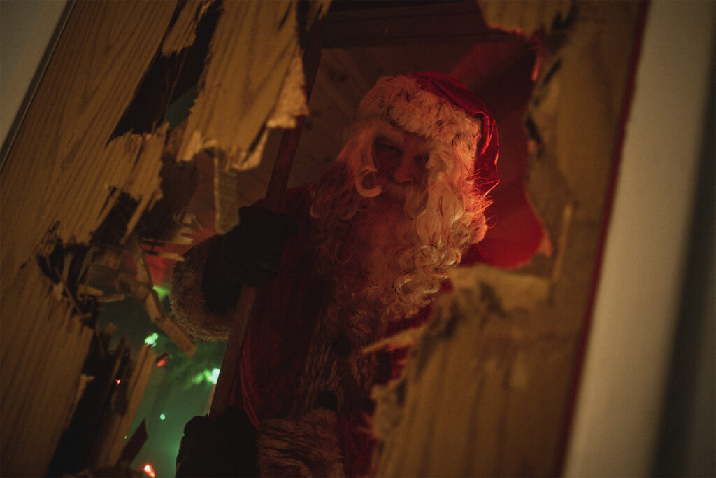 Image of Santa Claus covered in blood standing behind a broken down door in a scene paying Homage to Stanley Kubrick's 1980 masterpiece The Shining. Green and red Christmas lights are seen dangling behind Santa. His eyes staring fierce in front of him. His beard and long white locks drenched in the blood of his victims.  