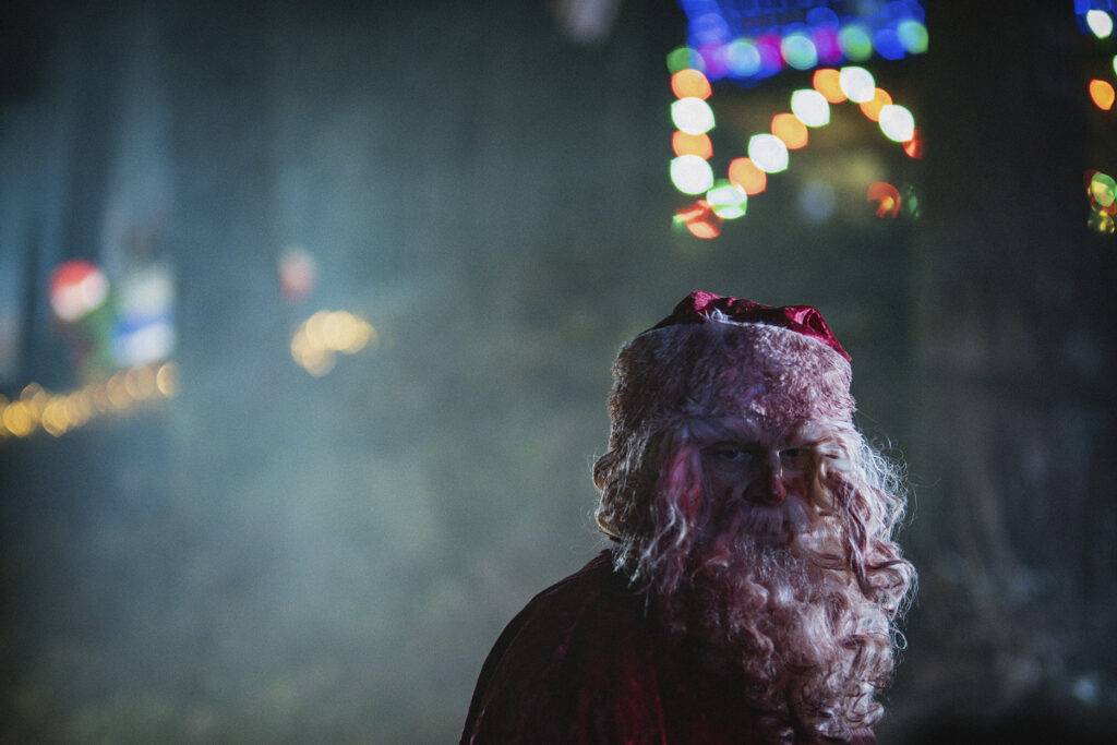 image of RoboSanta+ outside covered in blood. Crimson smears white curls in his bear and hair. His eyes emotionless. His iconic festive hat sits atop his head smeared in the same red liquid. Behind rests a festive light house blurred by fog. 