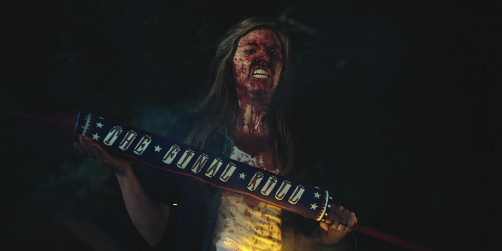 image of girl with blood splattered on her face running down her chin and neck, a snarling look of anger flooding over her. The background is hard to see as the lighting in the shot is really dark unfortunately. The character has long straight brown hair and is wearing a blue jean jacket over a bloody white shit. IN her hands she carries a festive looking club painted blue and red with stars (signifying the American flag) and the word The Final Kill reading down the side in bright red font. 