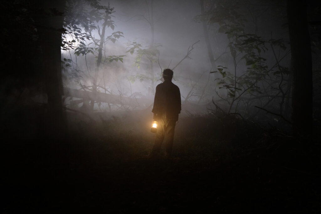 Dark image of Jos Ruben walingin a gloomy forested area. Fog encapsulates him and the screen as only the faint glow of his oil lamp helps lead his way. We see faints hints of green and browns, but the darkness eats away the light, leaving us feeling alone and isolated. 