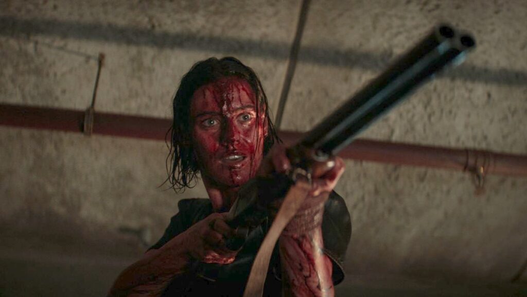 Closeup of Lilly Sullivan as Beth covered in blood. Her short brown hair wet and damp with the crimson liquid as it runs down her face and arms. In her hand she grips a double barrel shotgun pointed at someone or something off-screen. Her mouth slightly ajar as she begins to speak a line of dialogue. Above her head are rusted water pipes indicative of a basement setting. 