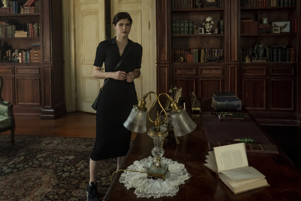Image of Alexandra Daddario as Rowan. Her long dark brown hair is pulled back as she stands with a look of curiosity on her face. Rowan is seen wearing a business casual black dress with a black leather purse draped around her left shoulder.  Her hands pressed together as she stands in a home study. Bookshelves filled with rare encyclopedias and other rare pieces of literature can be seen in the background. An old decadant lamp rest on a table in front of Rowan, an old book lying open, pages ready to flip on itself. 