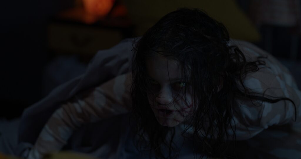 Image of Grace possessed. Blood streaks across her pale face as her long string like black hair shields her black demonic eyes. Her body is hunched over and covered in an orange and white striped shirt. 