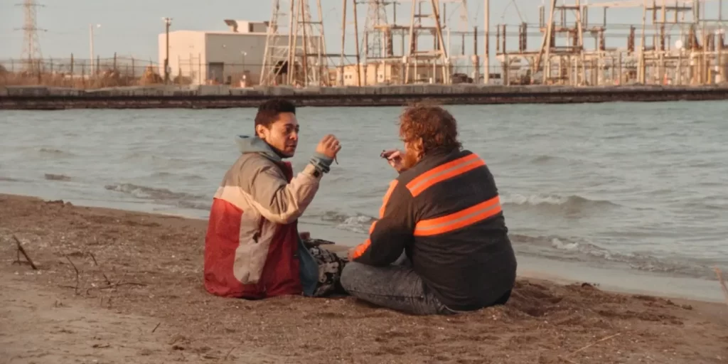actors Botello and Dawkins as their respective characters Roscoe and Benny sitting on a beach getting ready to eat a pair of worms together. Light waves can be seen rolling into the shore as Benny and Roscoe sit cross legged in the pebbled sand. Both are seen where light jackets. Roscoe is a combination of white, grey, and red colors while Benny's is black with two large orange strips running across his back, and one large orange strip along the side of his arm. Electrical towers can be seen in the back ground as Benny's urly brown hair blows in the wind, Roscoe peering at Benny with a look of anticipation. 