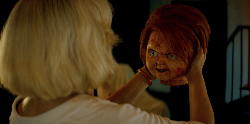 Jennifer Tilly as Tiffany Valentine holding the living decapitated head of Chucky. We can only see the back of Tiffany and her short bob blonde hair cut as she cradles Chucky's head. Chucky is seen with blood splatters on his face as his bright orange hair falls over his left eye. He stares back at Tiffany angrily, a sinister look peering deep into Tiffany's eyes. 