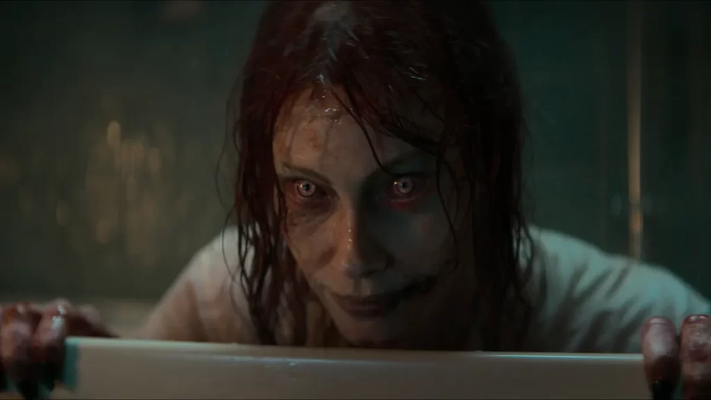 close up of Alyssa Sutherand sitting in a tub possessed as a Deadite. Her skin is pale and blotchy, thick sweat and plasma like substances runs down her face as her bright yellow demonic eyes stare into the camera. A light grin is on her face. Her hair damp and stringy adding to the sinister tone. 