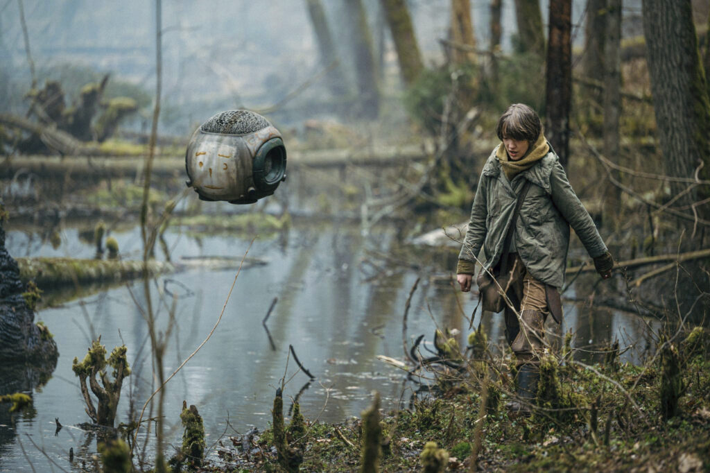 Vesper and her Robot walking through a marsh. Vesper is wearing brown pants, a large grey/green jacket and mustard yellow scarf draped across her neck. Her robot is seen with a face drawn on with what appears to be dry blood. Light mist fades in the background of the wooded marsh. 