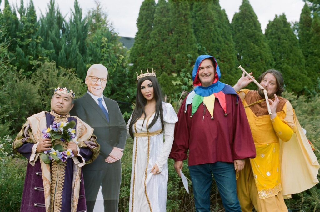 Picture of Joe Bob Briggs, Darcy the Mail Girl, John Patrick Brennan and Yuki Nakamura standing together dressed in medieval costumes. A cardboard cutout of Tom Atkins stands between Darcy and Yuki. Darcy is seen drapped in a beautfiul elegant princess dress, satin white with gold trim. Yuki is seen holding a small wreath of purple, white, and yellow flowers that match his loud medieval king costume. Resting atop both their heads are golden crowns. Joe Bob Briggs is seen standing to the left of Darcy, as he smiles whilst wearing a half-put together jester costumer. Lastly, we see Brennan with two wooden recorders in his hand as he mimics playing them both dress clad in a bright yellow dress. 