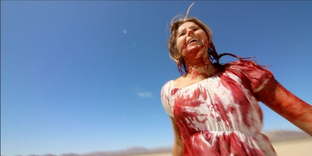 Actress Michelle May running manic in an open desert. She is dressed in a bloodied white summer dress, her hair sopping with the same crimson liquid covering her dress; blood smeared across her face and exposed arms. A bright blue cloudless sky hangs over head.