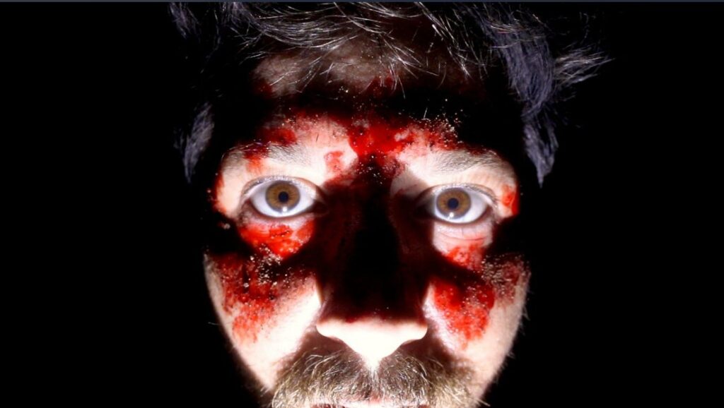 close-up shot of Writer/Direct/Actor Robbie Banfitch in pitch blackness. His face illuminate by his camera light. We see blood covering his disoriented face. 