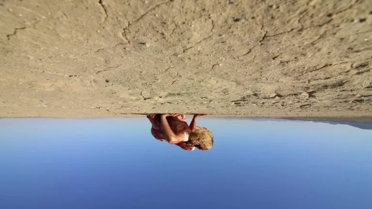 Disorienting upside down shot of Michelle lying in the desert. She is seen wearing the same blood covered dress, her face covered by her messy blonde and crimson caked hair. Her back points towards an inverted sky as the dry cracked desert floor lingers above. 