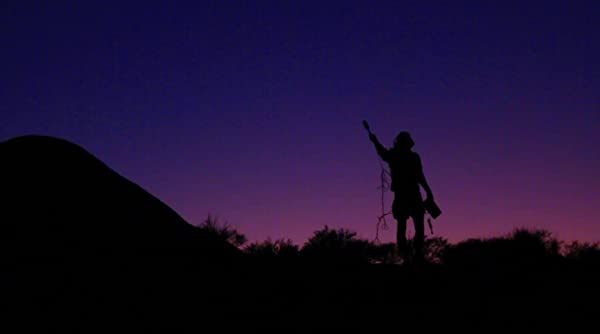 Beautiful wide shot of Robbie's silhouette standing atop a desert hill. His arm is extended up with his boom mic capturing the haunting sounds of the desert night. Hues of deep purples and midnight blues paint the sky above.  