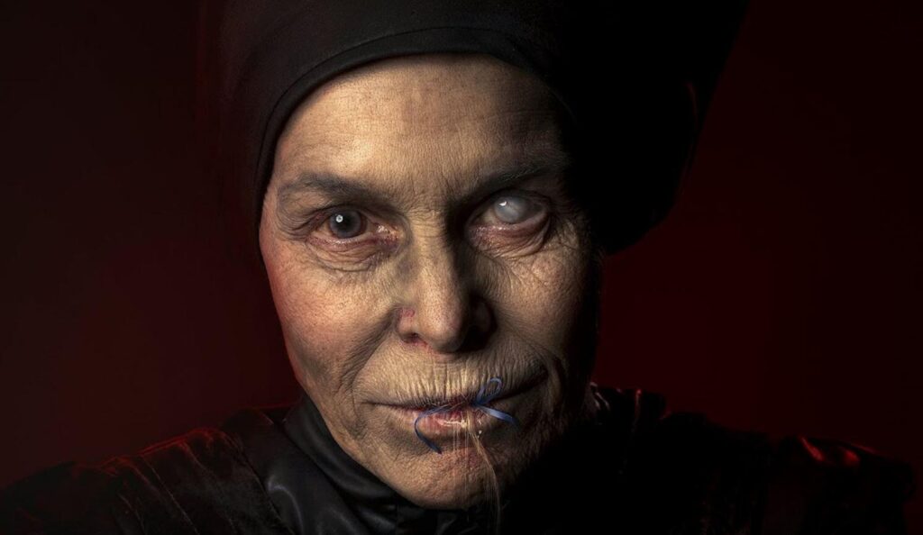 image of Alice Krige as The Witch in Gretel and Hansel. She stares in the camera with a sinister smirk. Her head covered and body drapped in black clothing. Her right eye a dark grey/brown color with her left pale white...blind. 