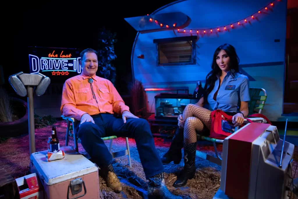 picture of Joe Bob Briggs and Darcy the Mail Girl sitting in lawn chairs on the set of The Last Drive-In. Behind them we see a white camping trailer with red string lights along the top and a bright red and blue neon sign that reads "The Last Drive-In' in the background. Joe Bob is seen wearing an orange collard button down long sleeve shirt and black jeans with snake skin cowboy boots. His hands rest on his thighs as he invites you in with a warm smile. Darcy is seen sitting to Joe Bob's left wearing a mail girl costume with fishnet stockings, her legs neatly crossed over one another. Surrounding the two hosts is an ice box with empty beer bottles and cans resting on the top and an old tube screen television in the front of them. 