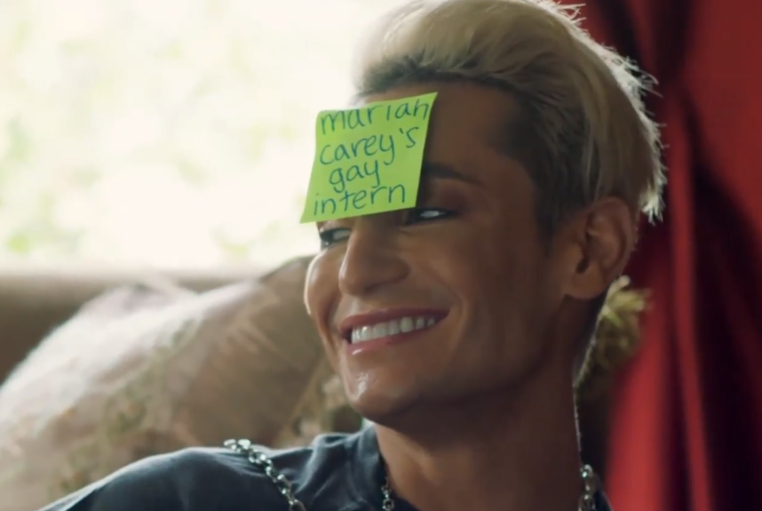 a party game scene where Nico is wearing a sticky note on his forehead that he can't read that says 'Mariah Carey's gay intern'