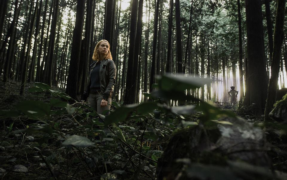 A girl in the forest during the day. She looks lost and confused. A creature lingers behind and away