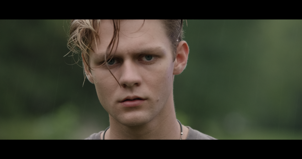 Ty Simpkins with wet hair, looking worriedly into the camera with a forest background