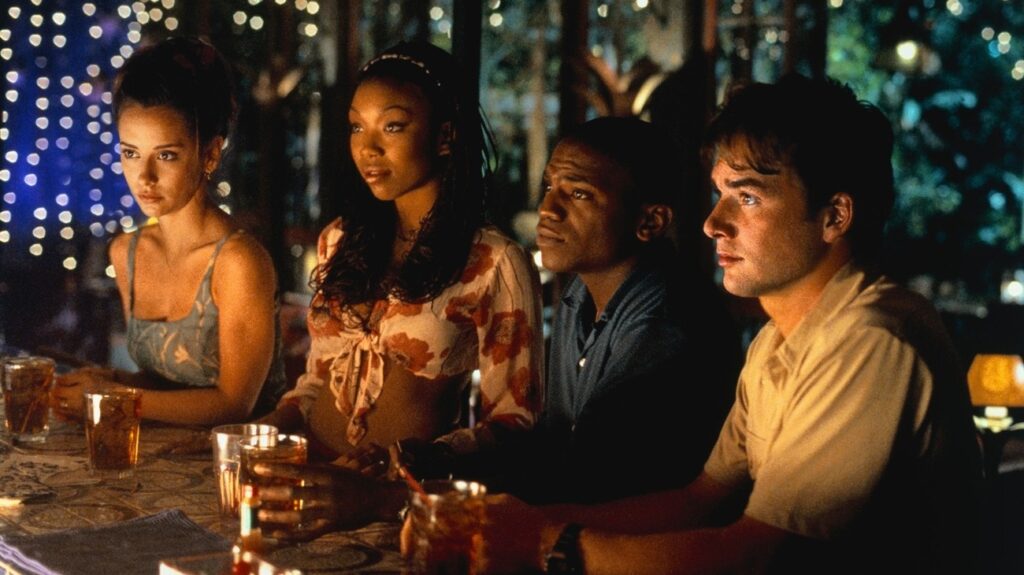 Four main characters are all dressed up in party clothing leaning on the bar looking at someone off camera.