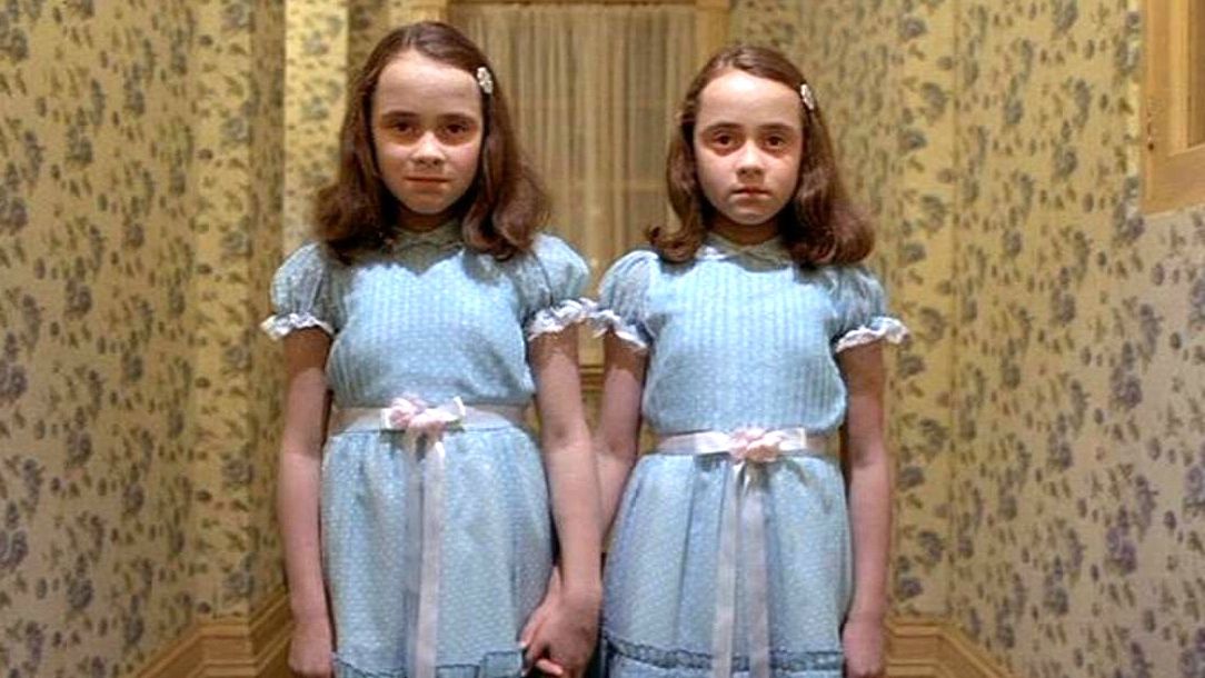 Two twins in blue dresses are standing against a yellow wall looking menacing