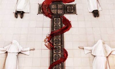 Nuns laying on a tile floor, possing in a T shape. Between them is a cross with a red painted serpent wrapped around it.