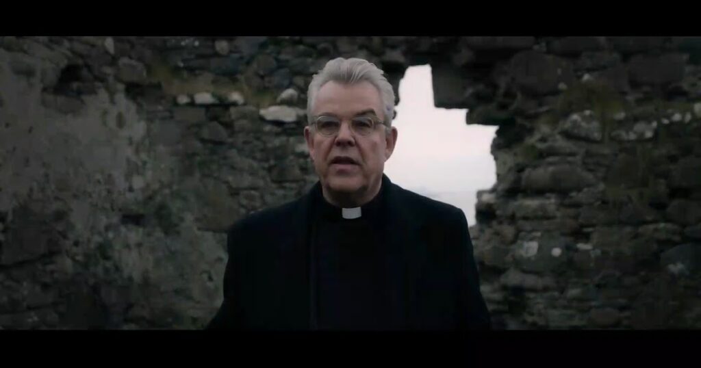 A priest standing in front of a stone structure. In the middle of the structure is an opening revealing a clear sky.