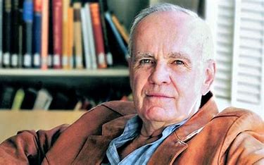 Cormac McCarthy sitting for an interview. A collection of books rest on a bookshelf behind him.