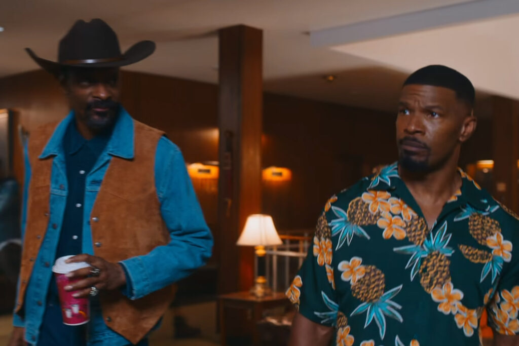 Man dressed as a cowboy and another man in a Hawaiin shirt walking in a building.