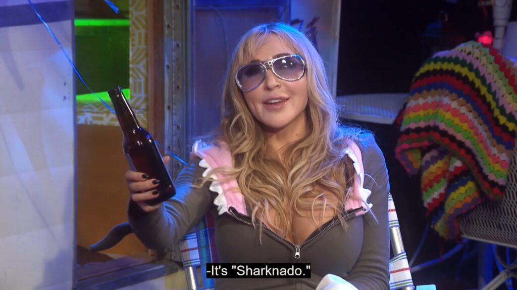 Darcy the Mailgirl, wearing a shark hoodie and sunglasses, gestures towards an off-screen Joe Bob Briggs with a beer bottle. The caption on the image reads "It's Sharknado."