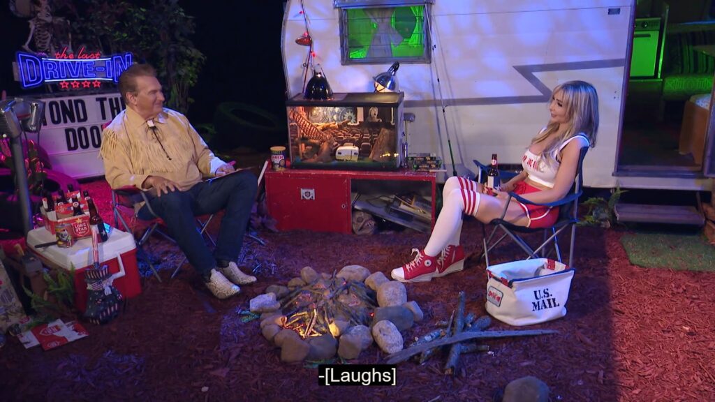 Joe Bob and Darcy share drinks and laughs while sitting in front of a fake campfire.
