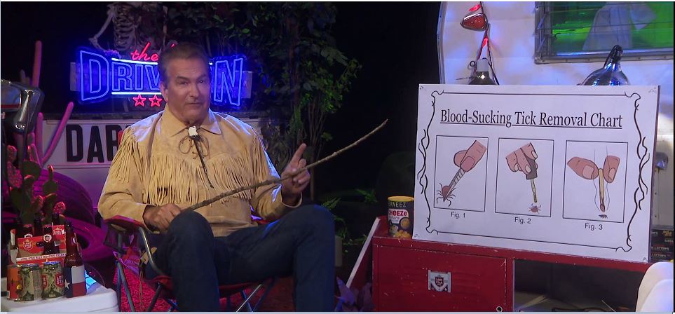 Joe Bob Briggs sits in a camping chair on the folk horror version of the trailer park set. He is holding a stick and is next to a poster which reads "Blood-Sucking Tick Removal Chart." The chart shows a tick being removed by tweezers, a tick being suffocated with nail polish, and a tick being burnt with a match.