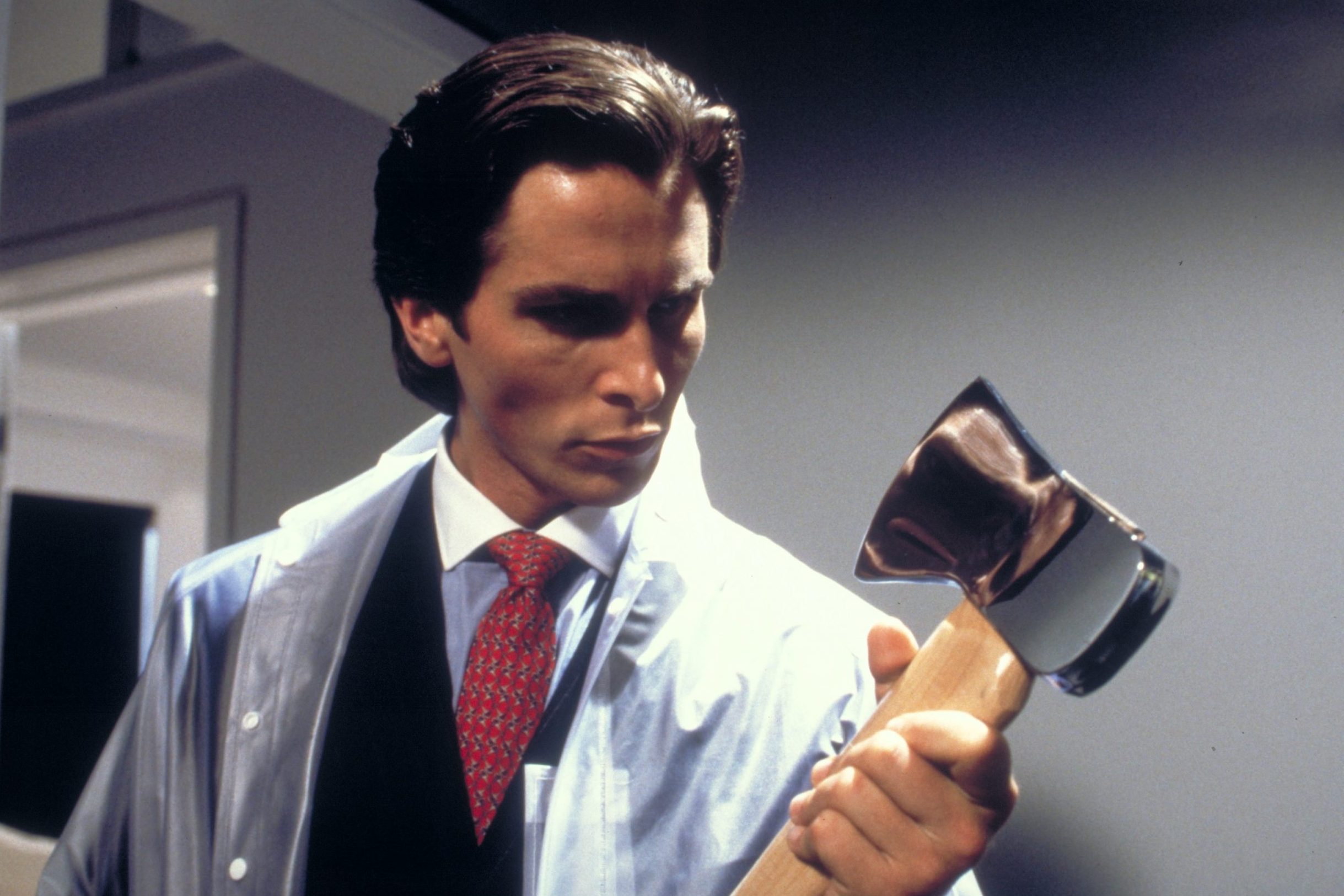 Patrick Bateman is holding an axe looking down on it while holding it in his arms. He's wearing a seethrough coat on top of a business suit.